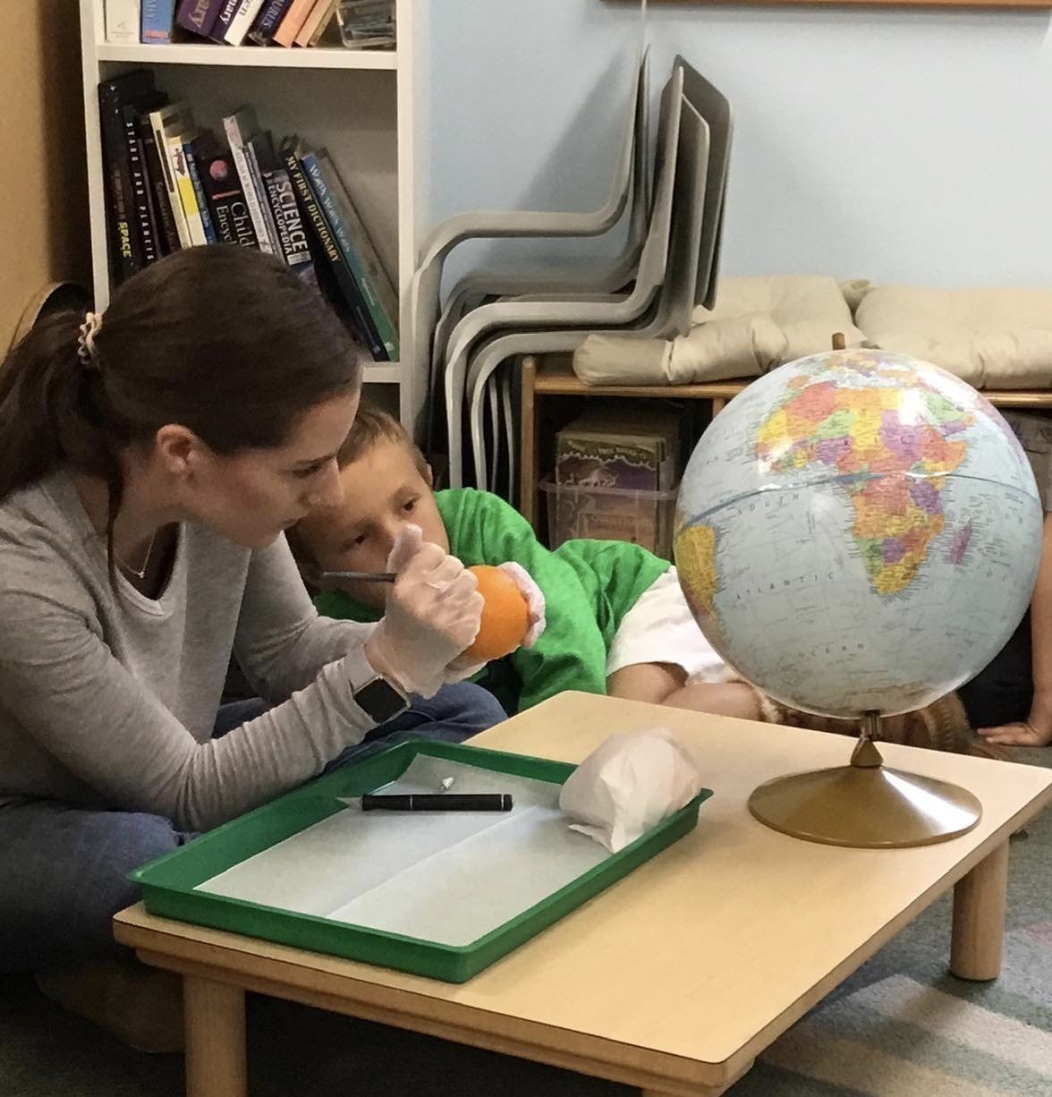 A montessori guide sits on the floor with a child and a globe on a table