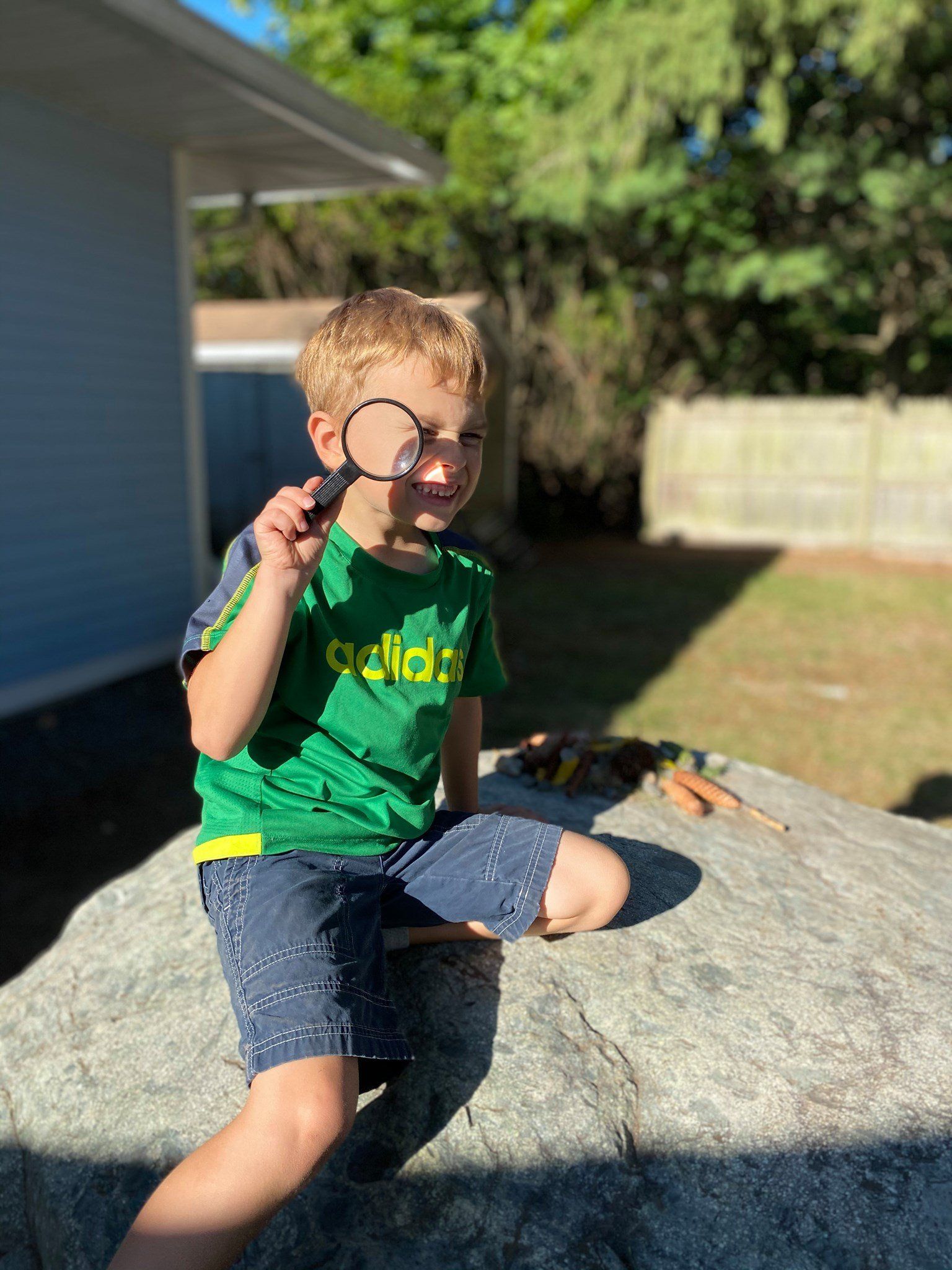 A young boy is sitting on a rock looking through a magnifying glass.