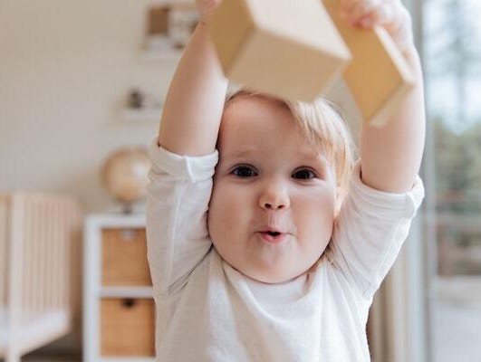 Montessori toddler raising wooden blocks, smiling with arms up. Indoor setting with a globe and shelves.