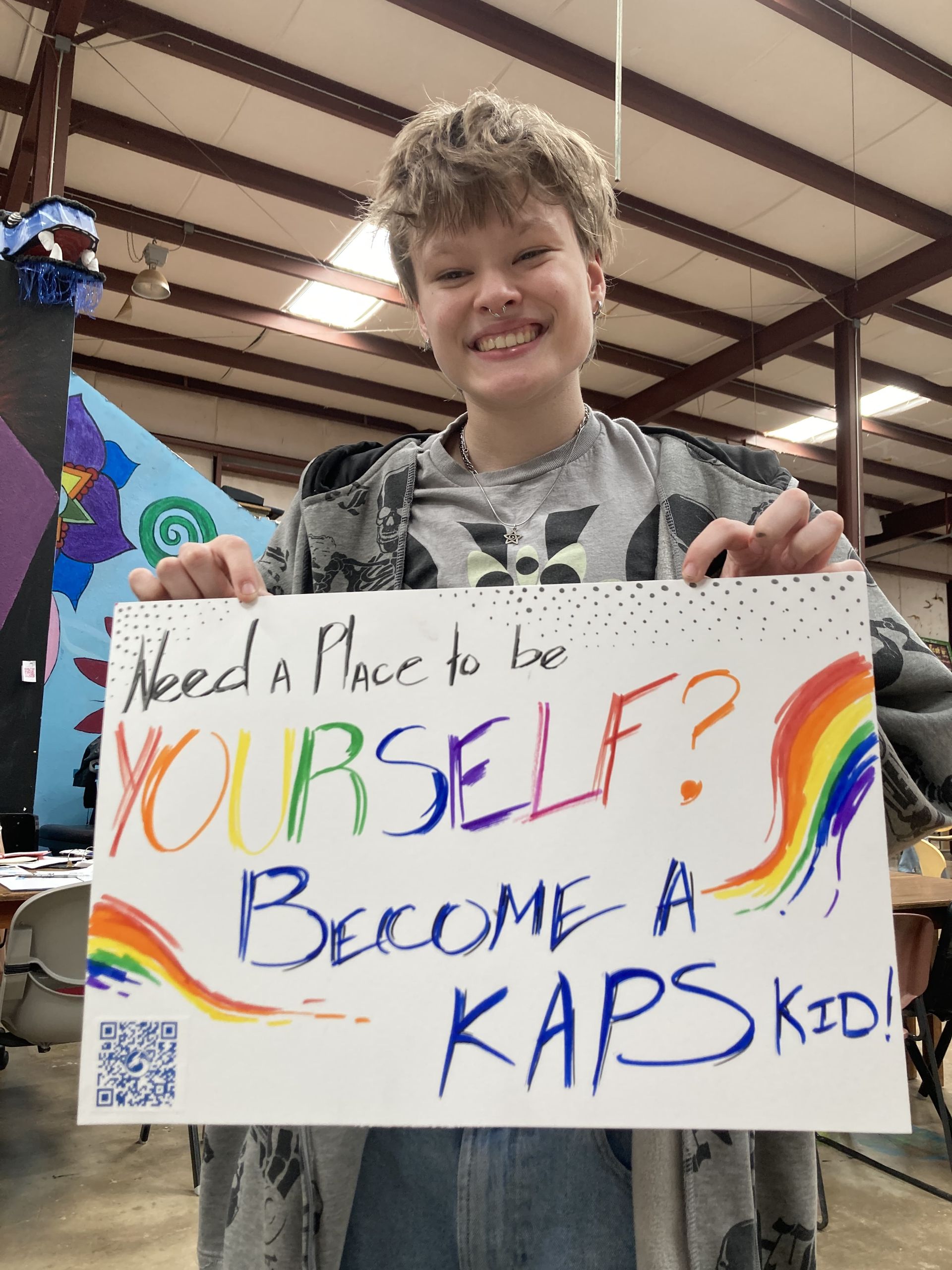 Frankie, holding a poster stating 'Need a place to be Yourself? Become a KAPS kid!'
