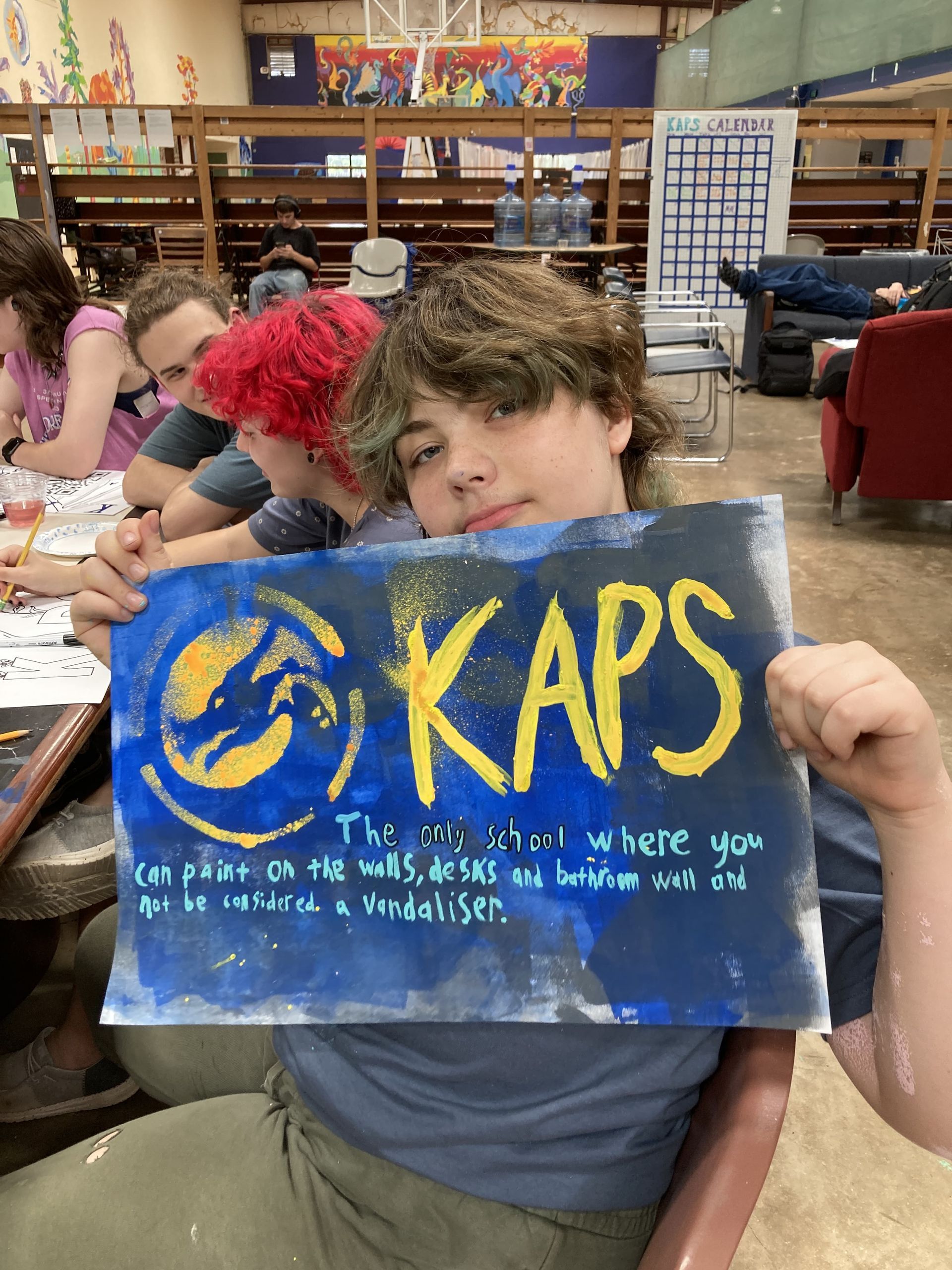 Spencer B., holding a poster saying 'KAPS - The only school where you can paint on the walls, desks and bathroom wall and not be considered a vandalizer.'