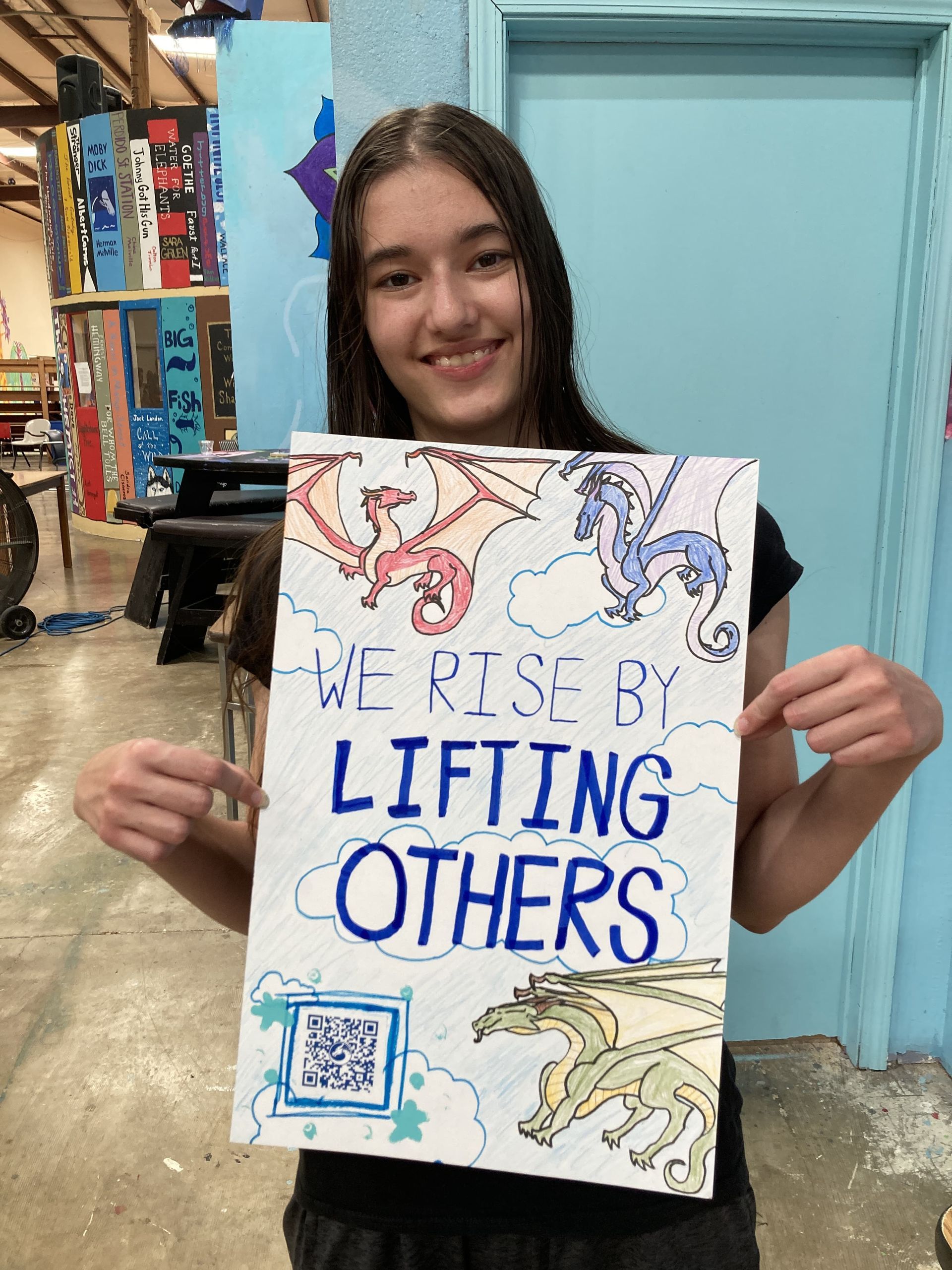 Celeste S., holding a poster that states 'We Rise by Lifting Others'