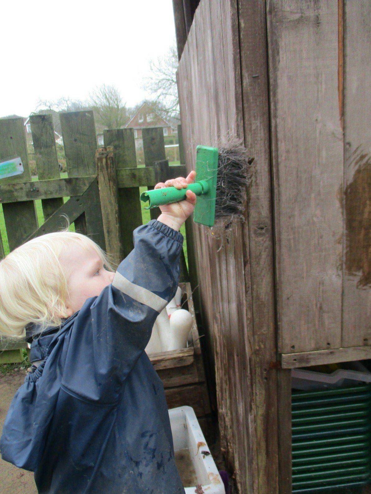 A little girl is painting a wooden wall with a green brush.