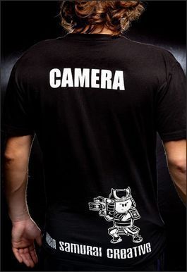 Man wearing a black shirt with "CAMERA" on the back and a cartoon samurai holding a camera.