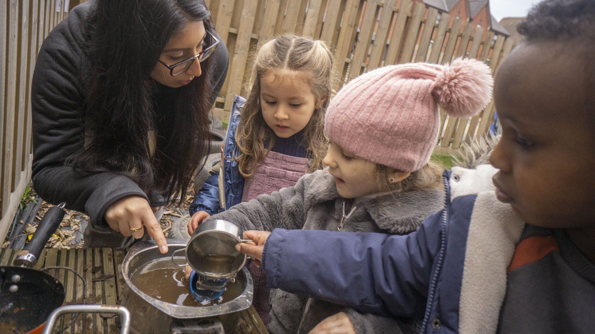 Teacher and children pouring liquid into a metal container outside near a wooden fence.