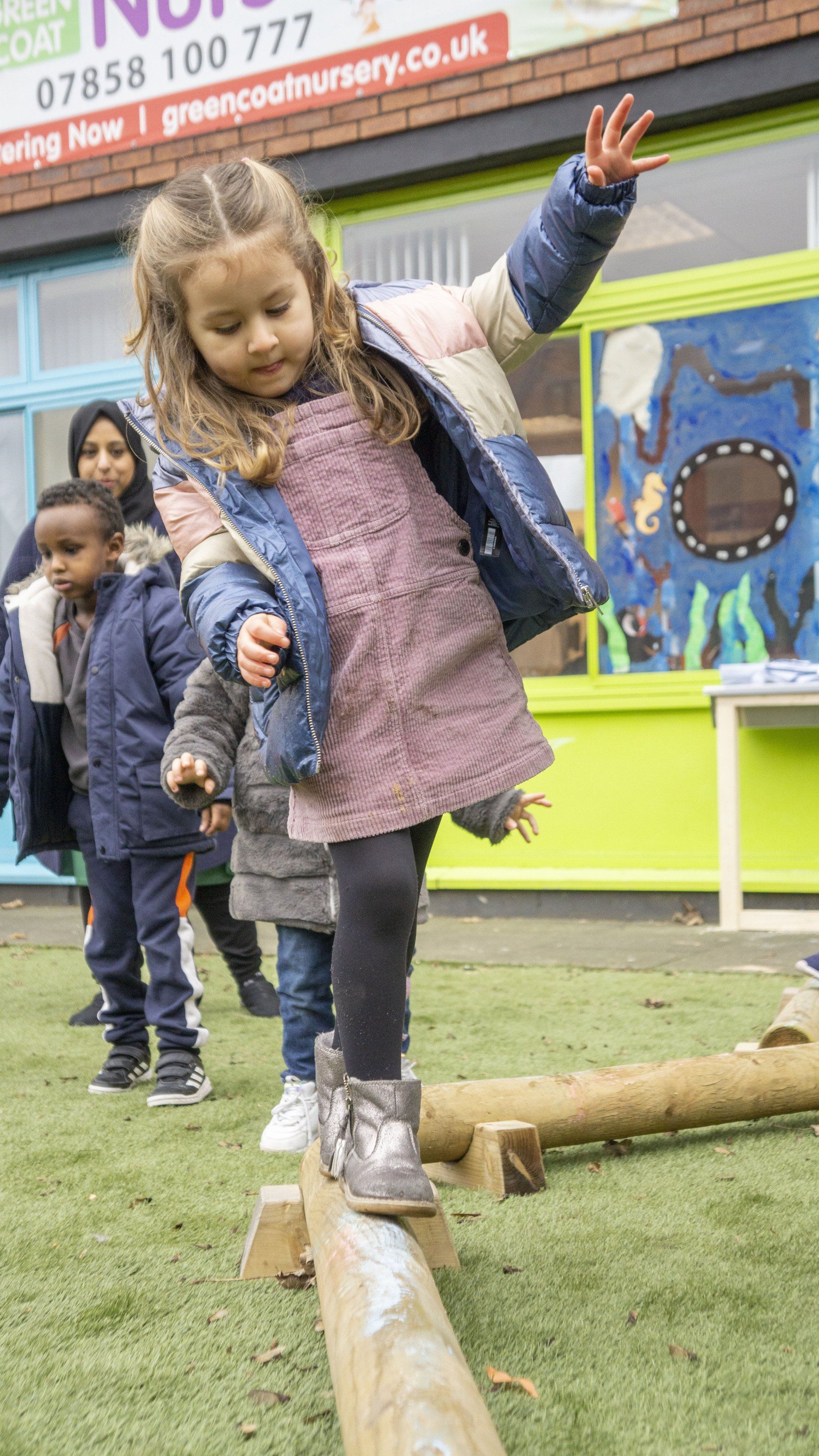 Girl walking on wooden beam outdoors, balancing, other children in background.