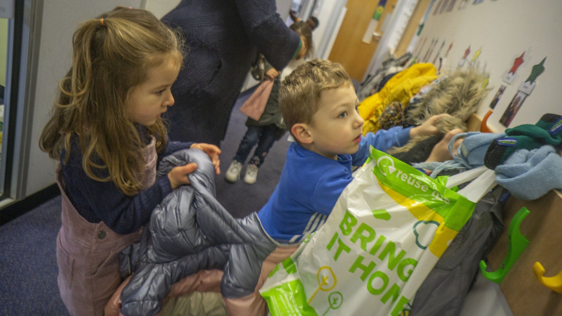 Girl and boy sorting through coats in a brightly lit hallway. A 