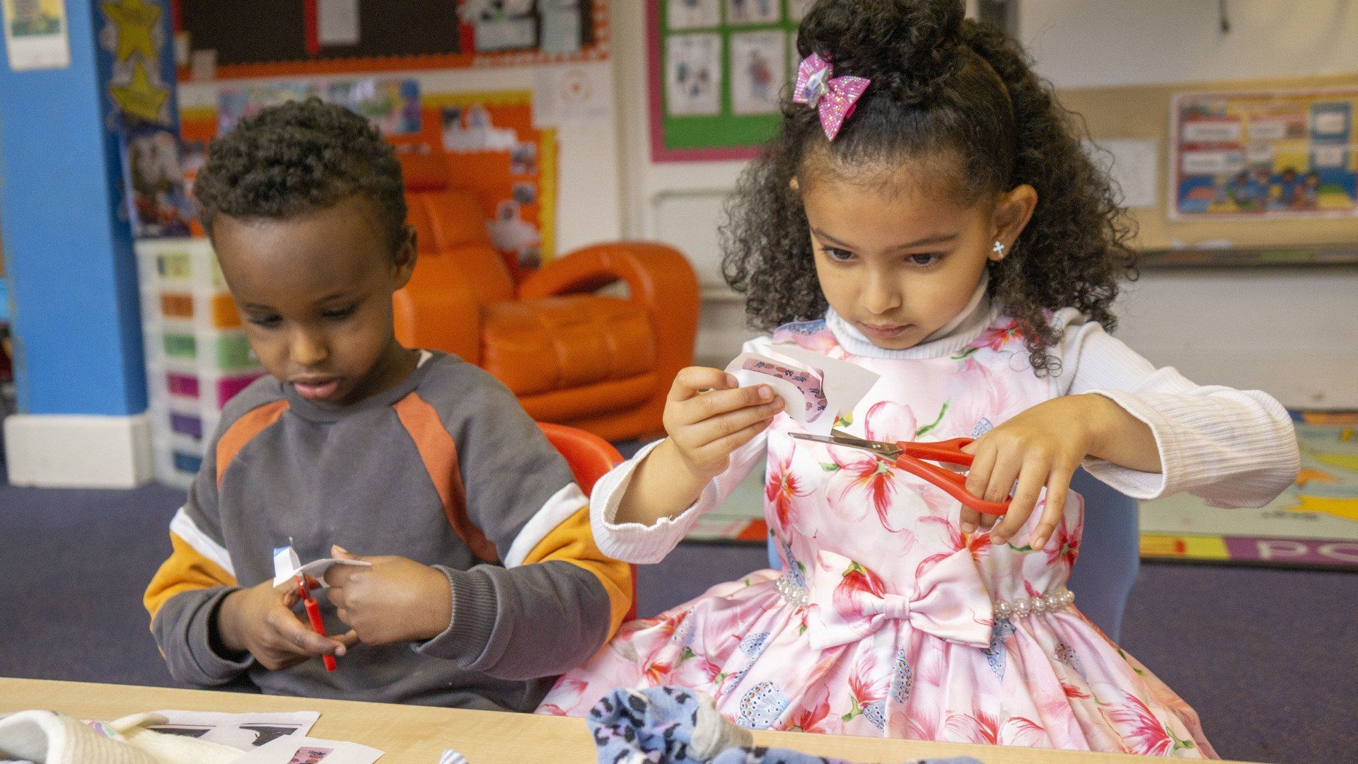 Two children cutting paper at a table in a classroom.
