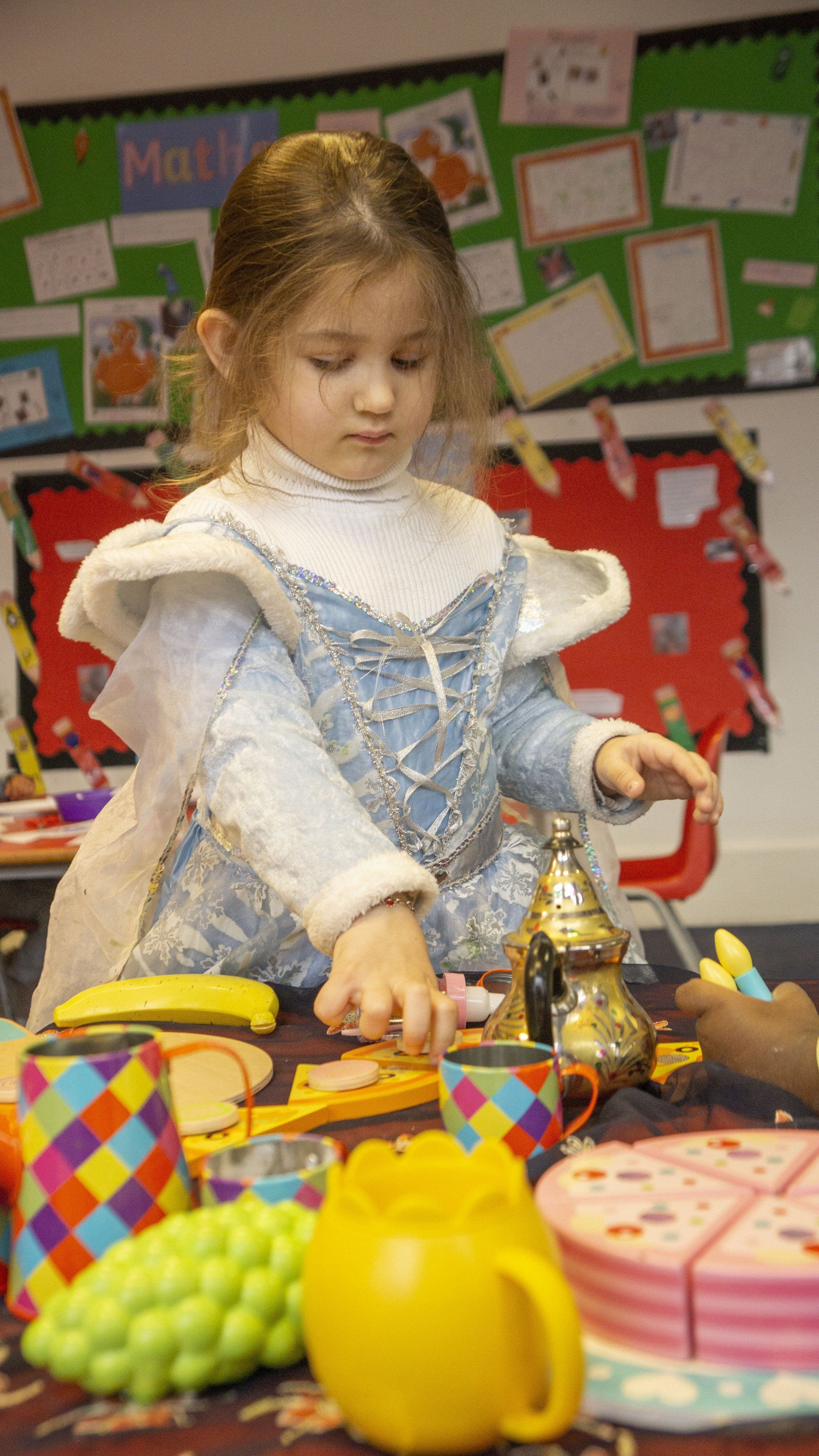 Girl in a blue dress playing with toy food in a classroom, focused and reaching.