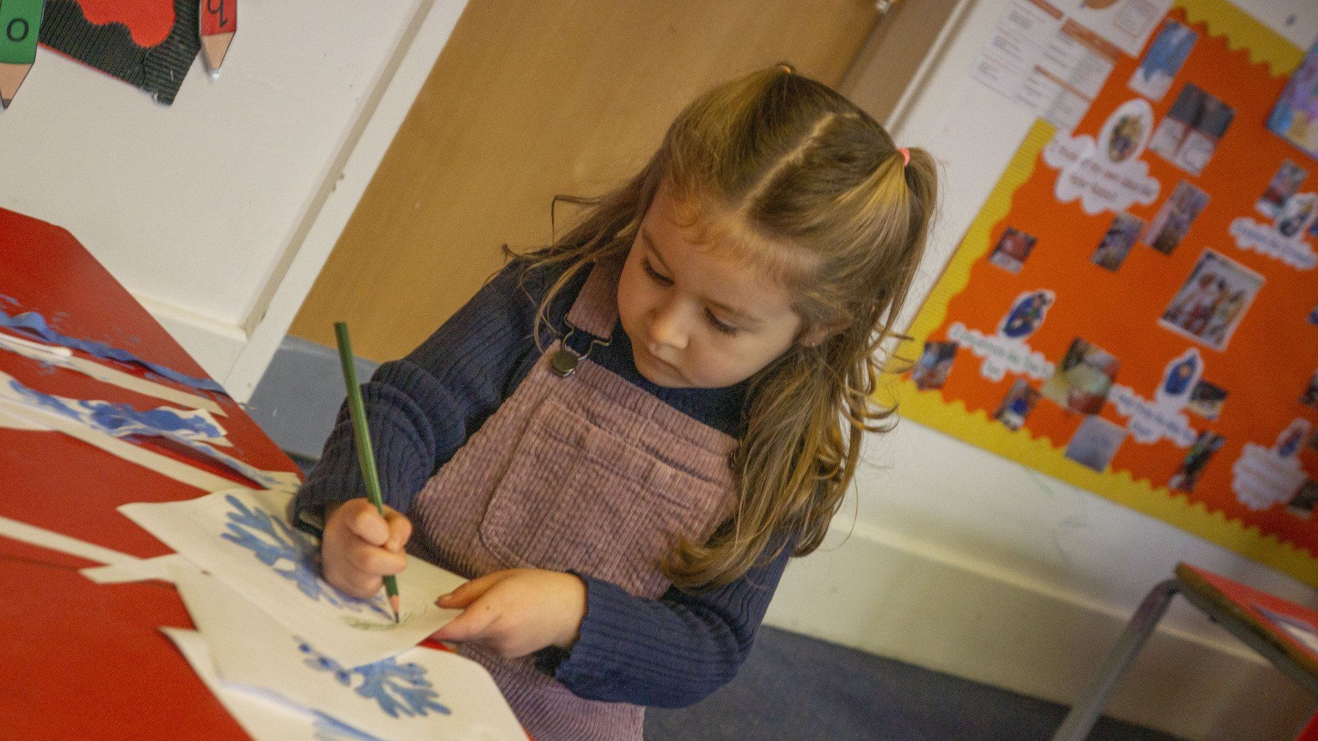 Young child with light brown hair coloring paper at a red table in a classroom.