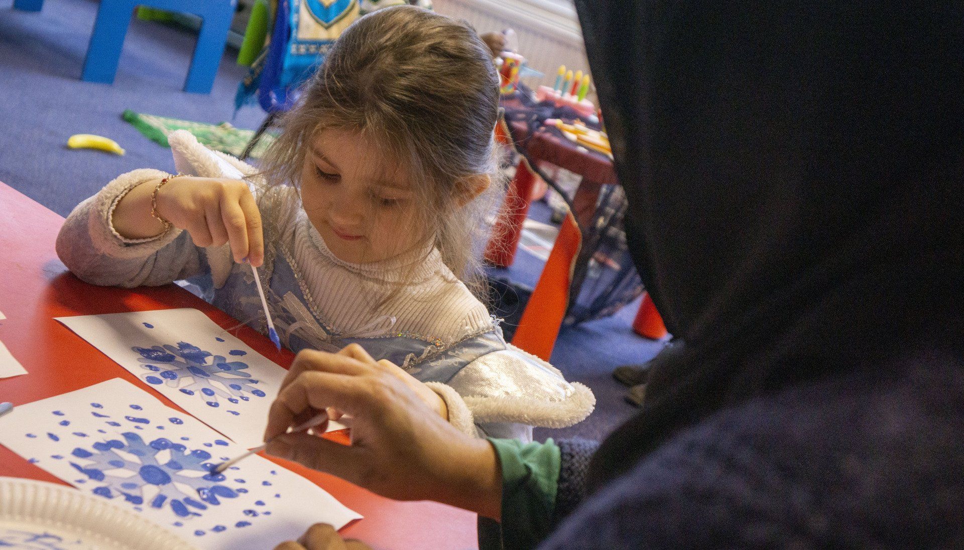 Girl painting snowflakes with assistance from a person in a head covering at a red table.