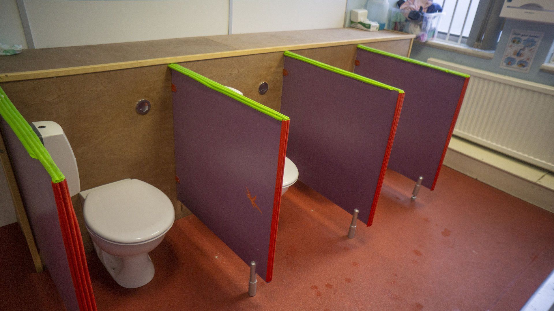 Three child-sized toilet stalls with purple doors, red trim, and green edges in a red-floored bathroom.