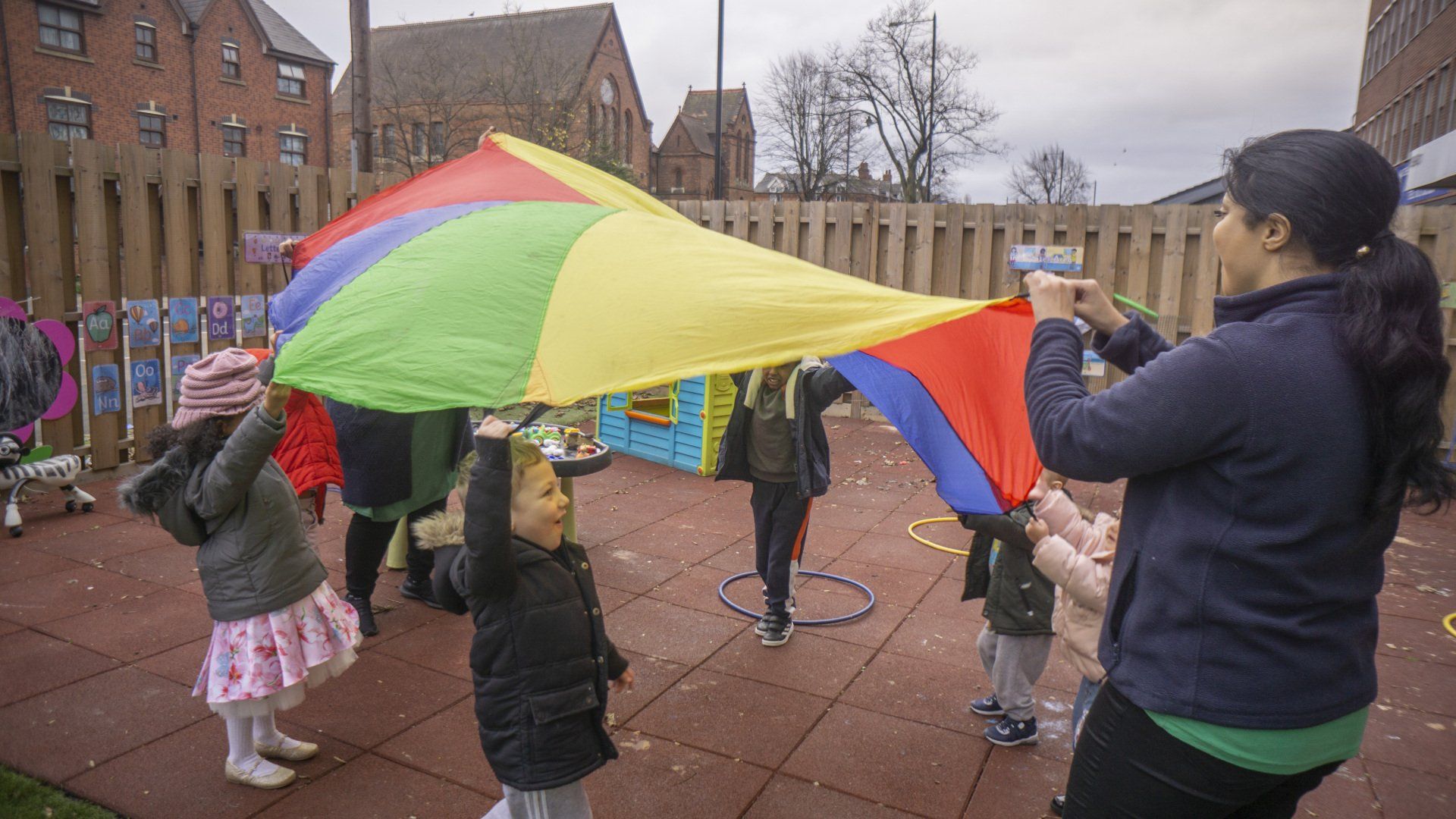 Children and an adult playing with a rainbow parachute in a playground.