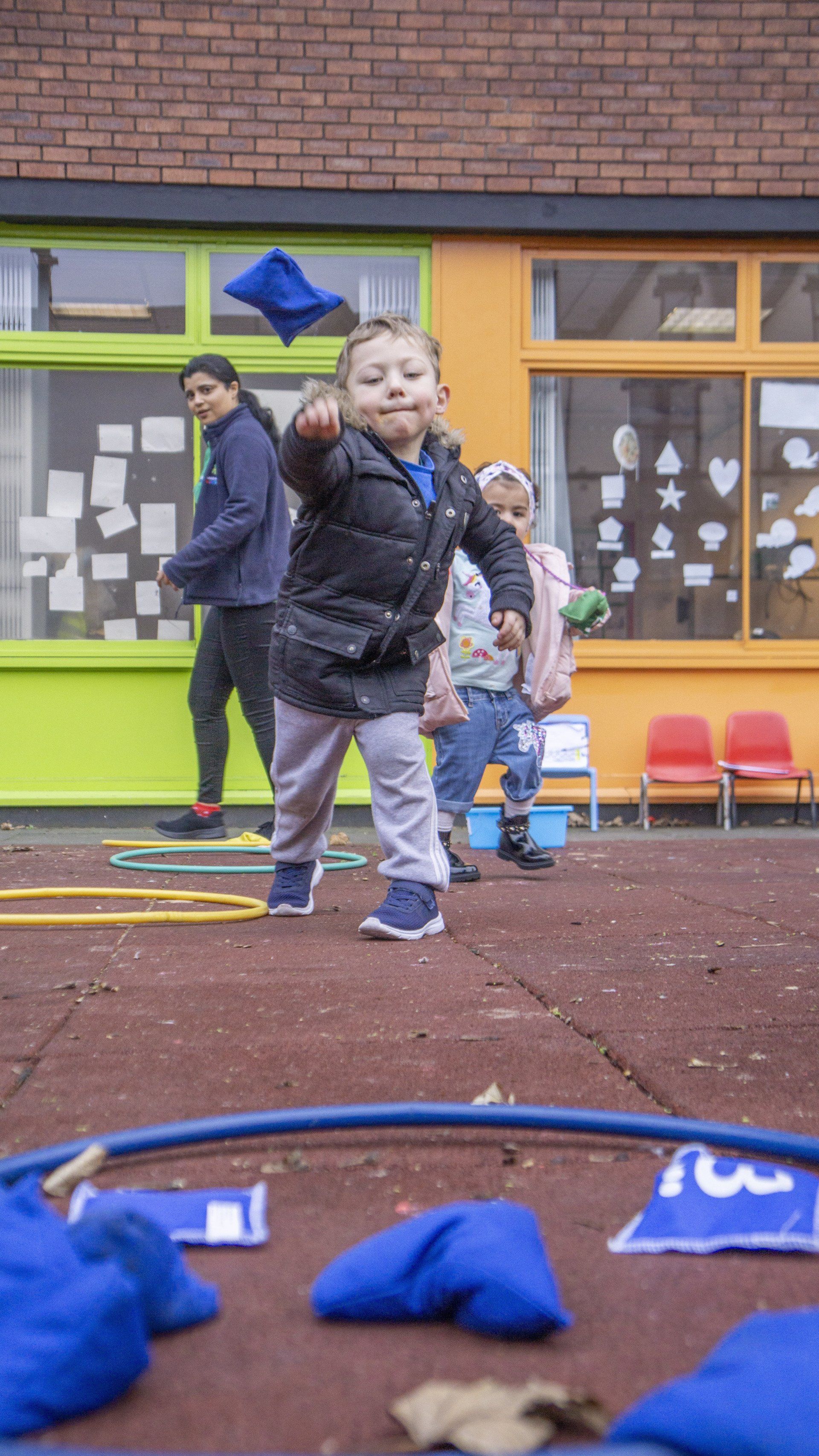 Child throws blue bean bag into a hoop during an outdoor game. Others watch.