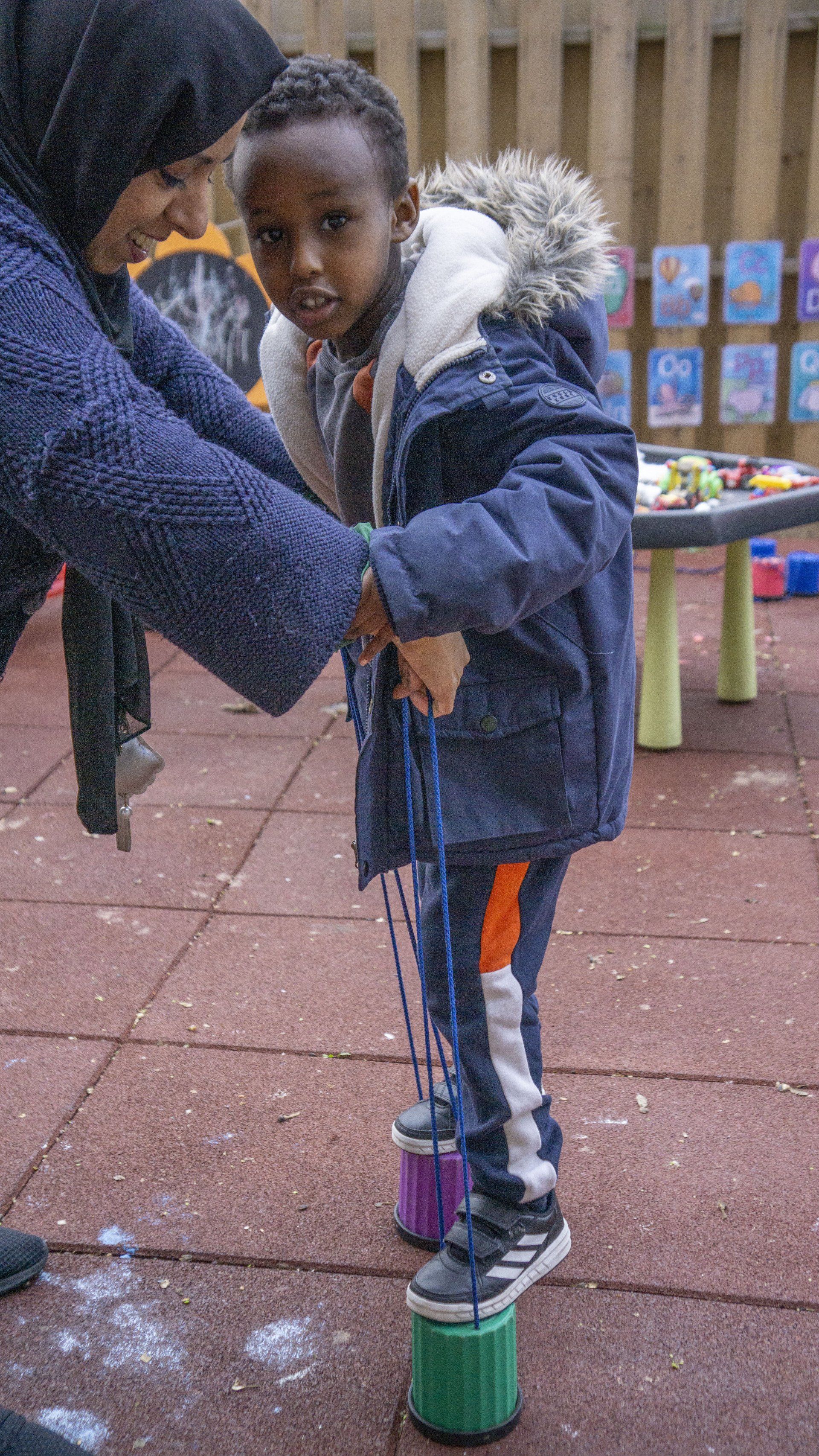 Child using stilts with assistance from a person outdoors, wearing winter clothes.