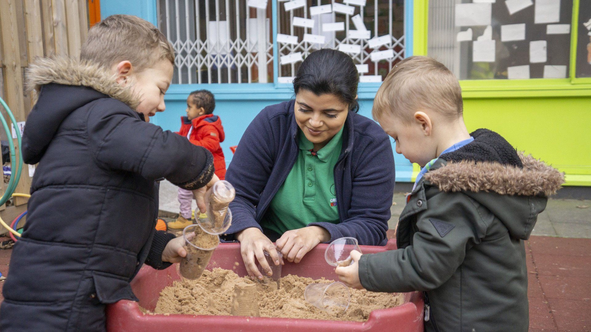 Children and an adult playing in a sand pit, outdoors. The adult is smiling.