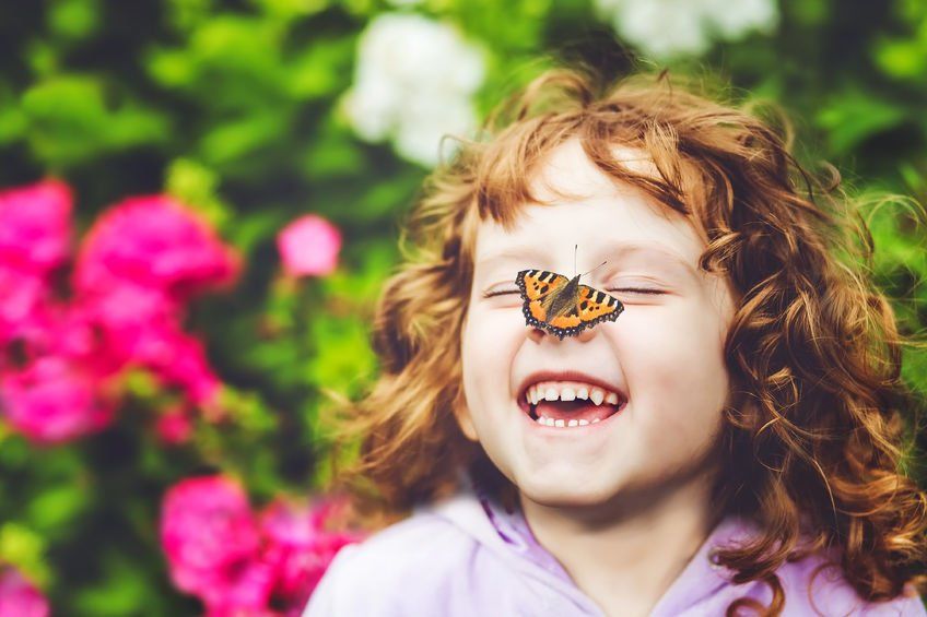 Girl smiling with a butterfly on her nose in a garden with pink and green flowers.