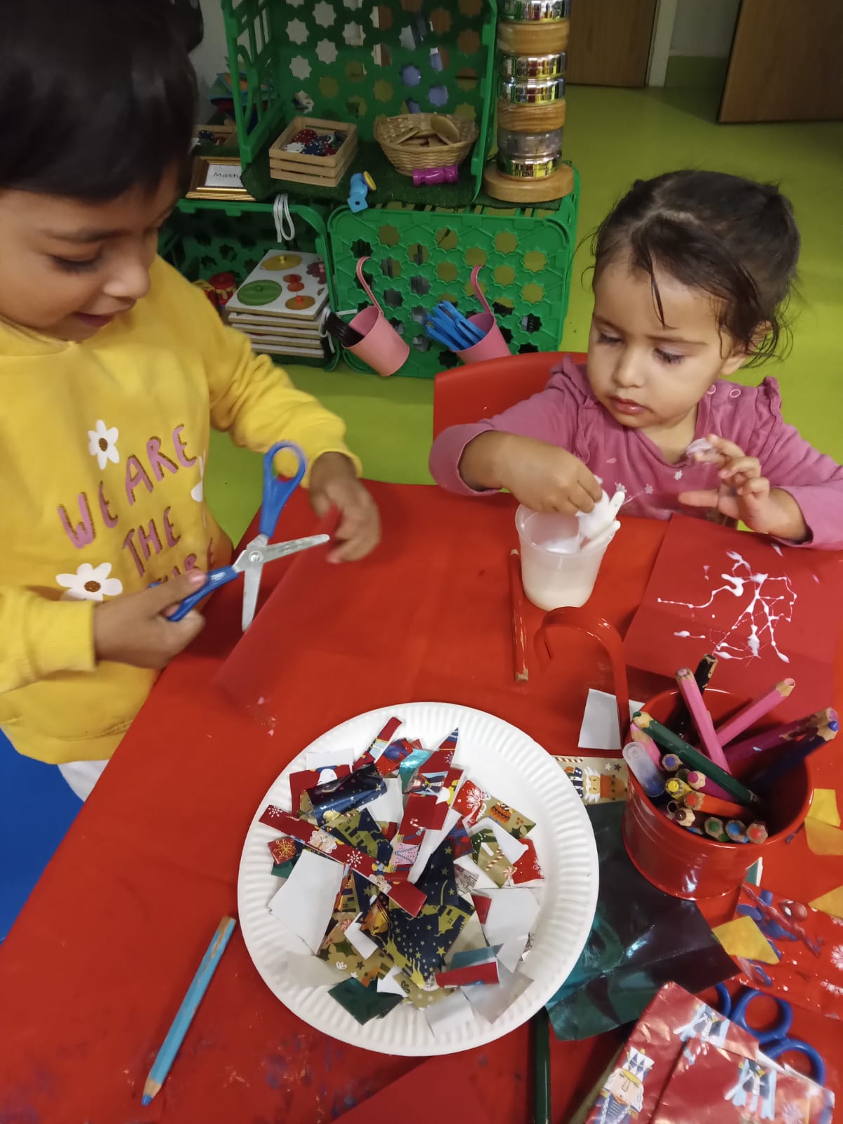 Two children crafting at a red table: one cuts paper with scissors, the other works with glue, surrounded by art supplies.