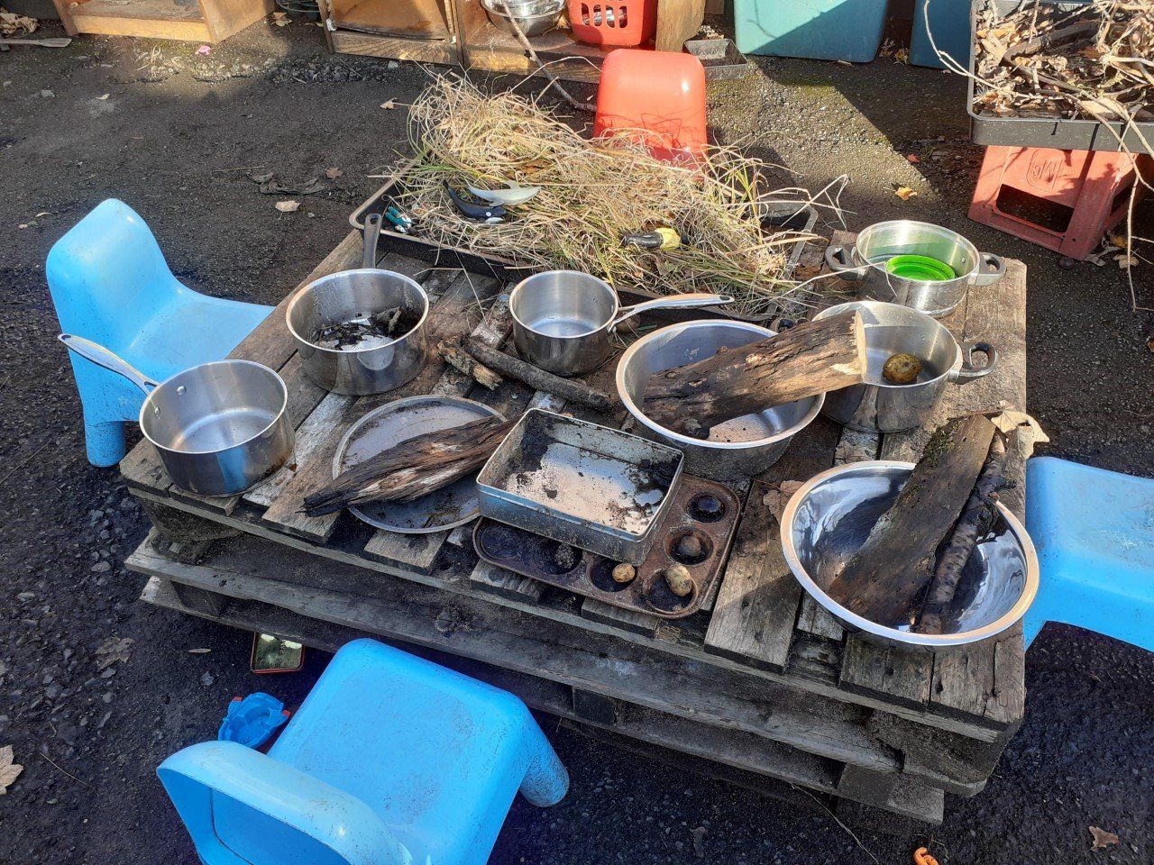 A low wooden pallet with pots, bowls, and materials for play, surrounded by blue plastic chairs.
