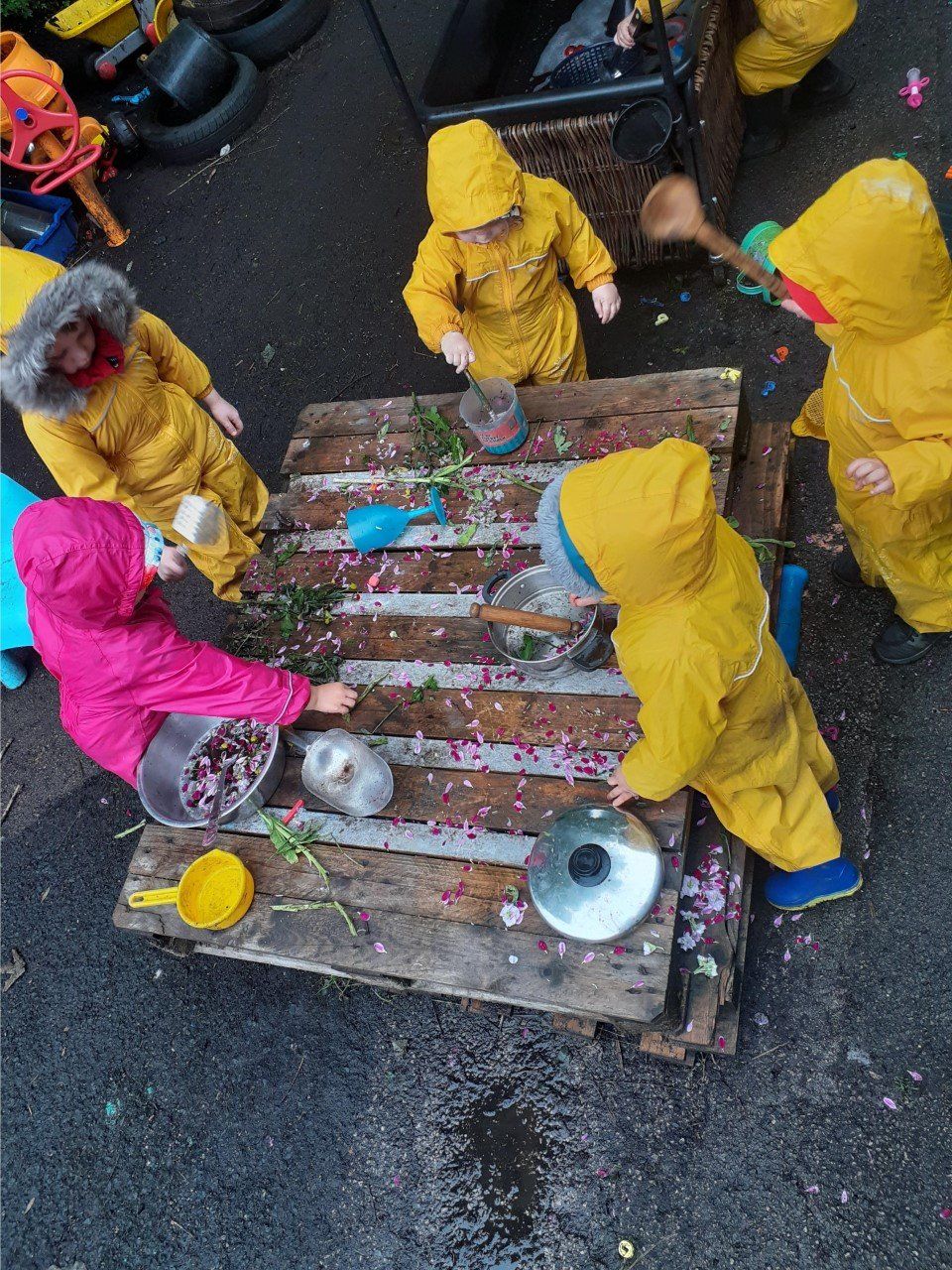 Children in yellow rain gear play at a wooden table with pots, pans, and flowers.