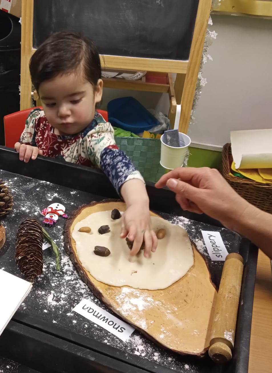 Child at a table, reaching for items on a wooden platter with flour, next to the word 