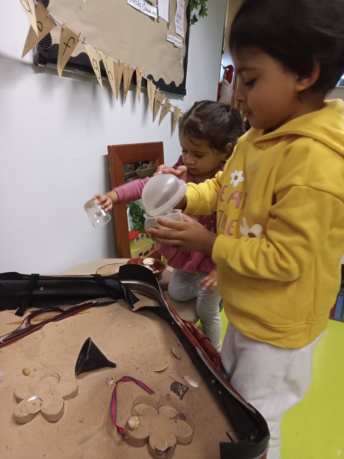 Two children pouring sand from cups at a sensory table. One child wears a yellow hoodie.