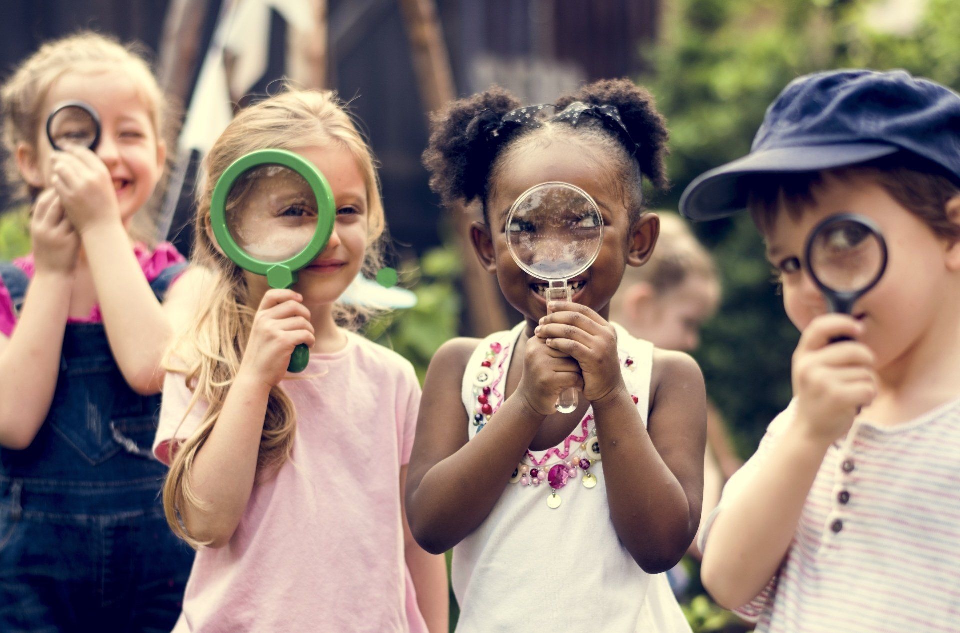 Four children looking through magnifying glasses. Outdoors, smiling.