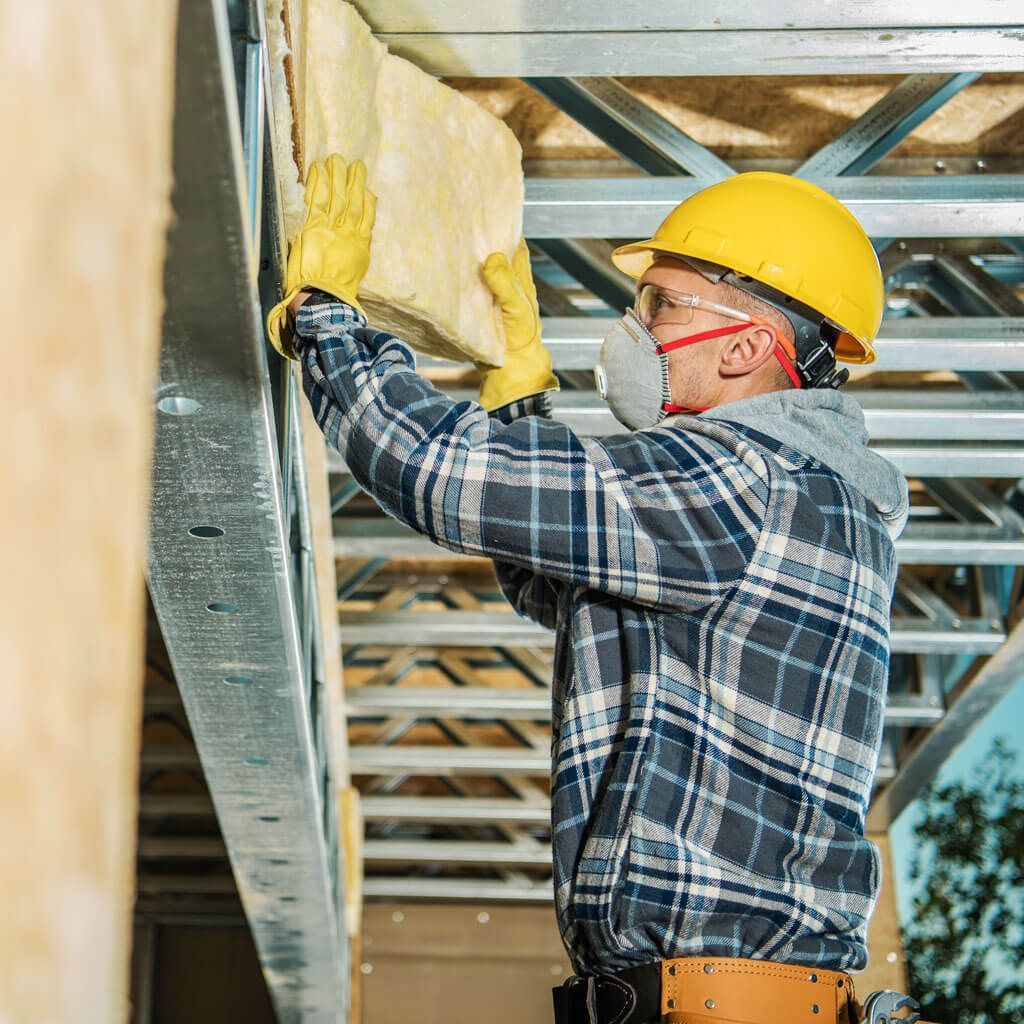 A construction worker wearing a hard hat , mask and gloves is insulating a wall.