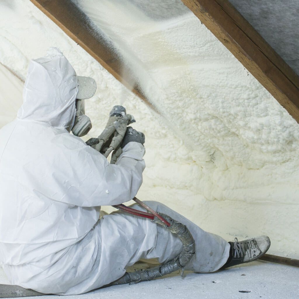 A man in a white suit is spraying insulation in an attic.