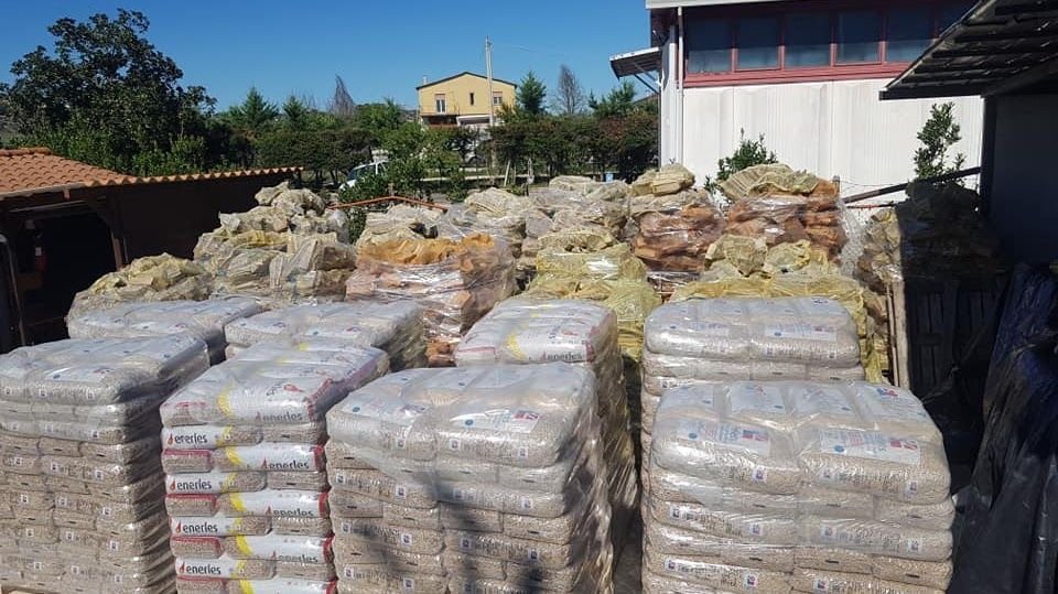Pile di pallet avvolti nella plastica, contenenti sacchi di pellet e altri materiali, si trovano all'aperto vicino a un edificio.