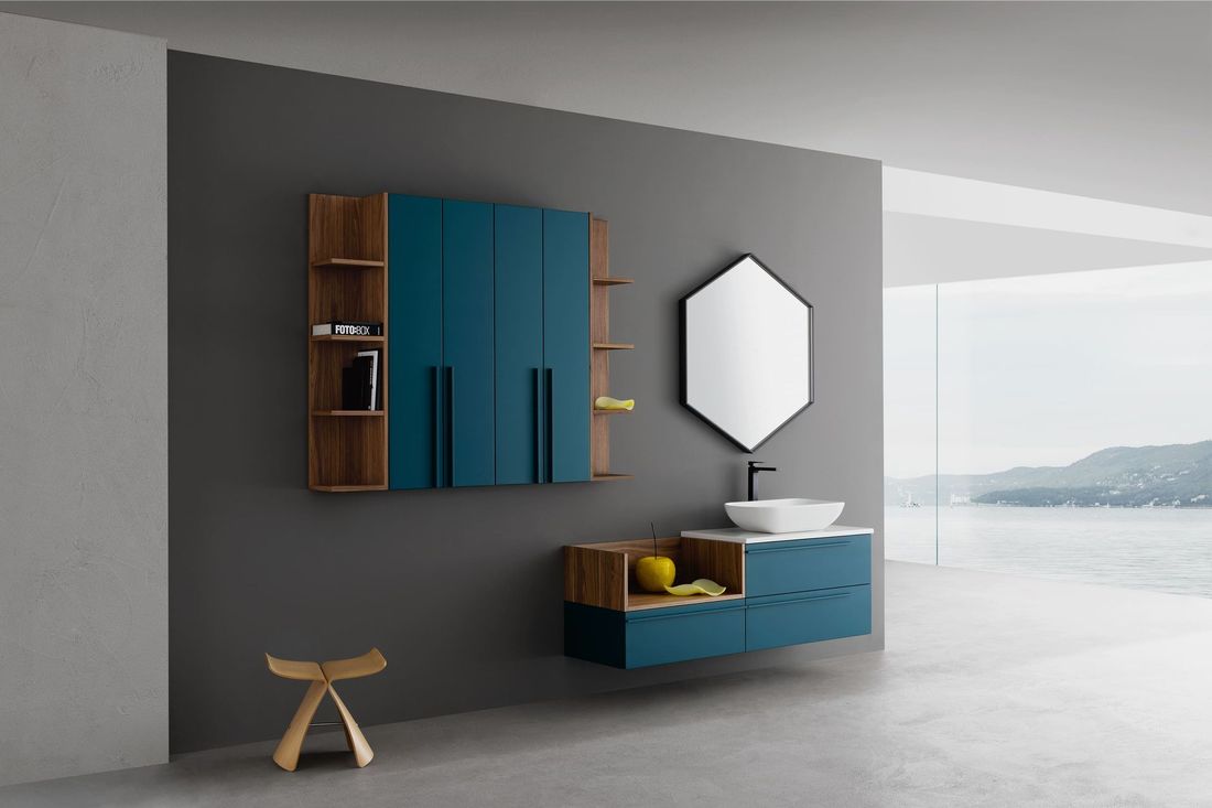 Camera moderna sui toni del grigio con pensili blu, specchio esagonale e una panca bassa accanto a una finestra luminosa.