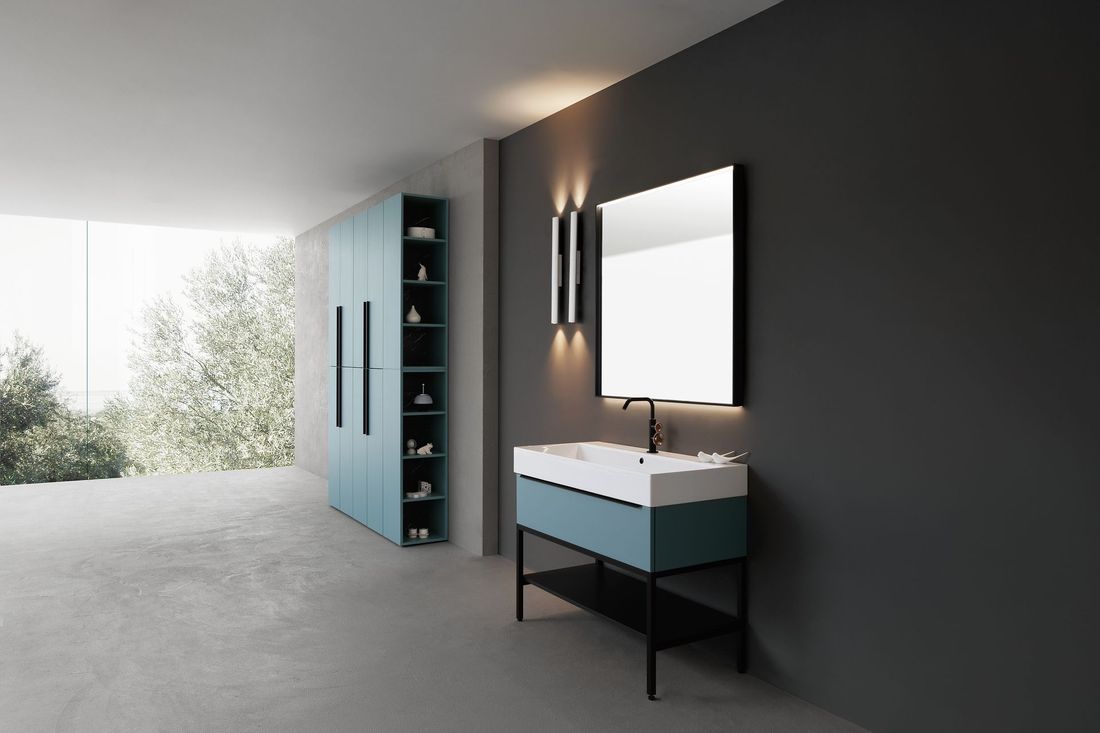 Bagno moderno con parete scura, mobile lavabo sospeso, luci a specchio e mobiletto blu accanto a una grande finestra.