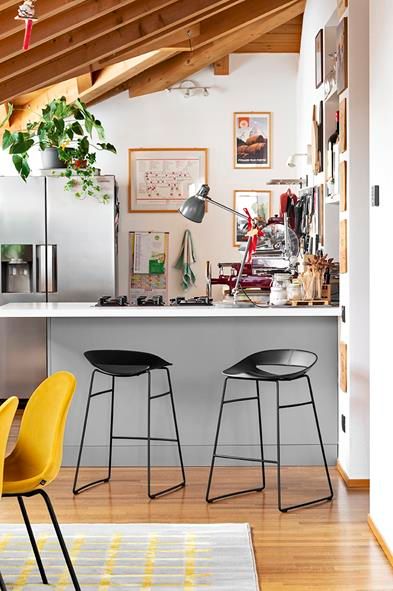 Cucina moderna con isola, due sgabelli neri, sedia gialla, piante e soffitto con travi in ​​legno a vista.