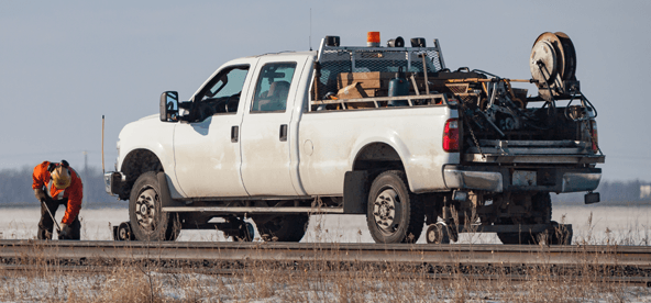 White truck on railroad tracks, worker in orange vest. Snowy field in background.