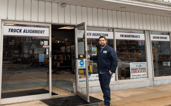 Man in blue uniform stands at the door of an auto repair shop with services listed in the window.