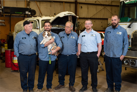 Five mechanics with a puppy in a garage. They are standing in front of a truck, smiling.