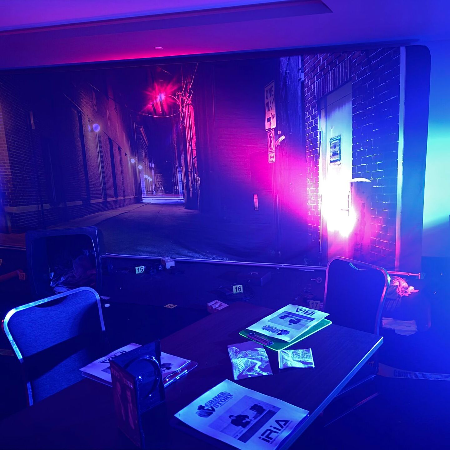 Table set up in front of a backdrop of an alleyway; with brochures and two chairs. Lit with blue and pink lights.