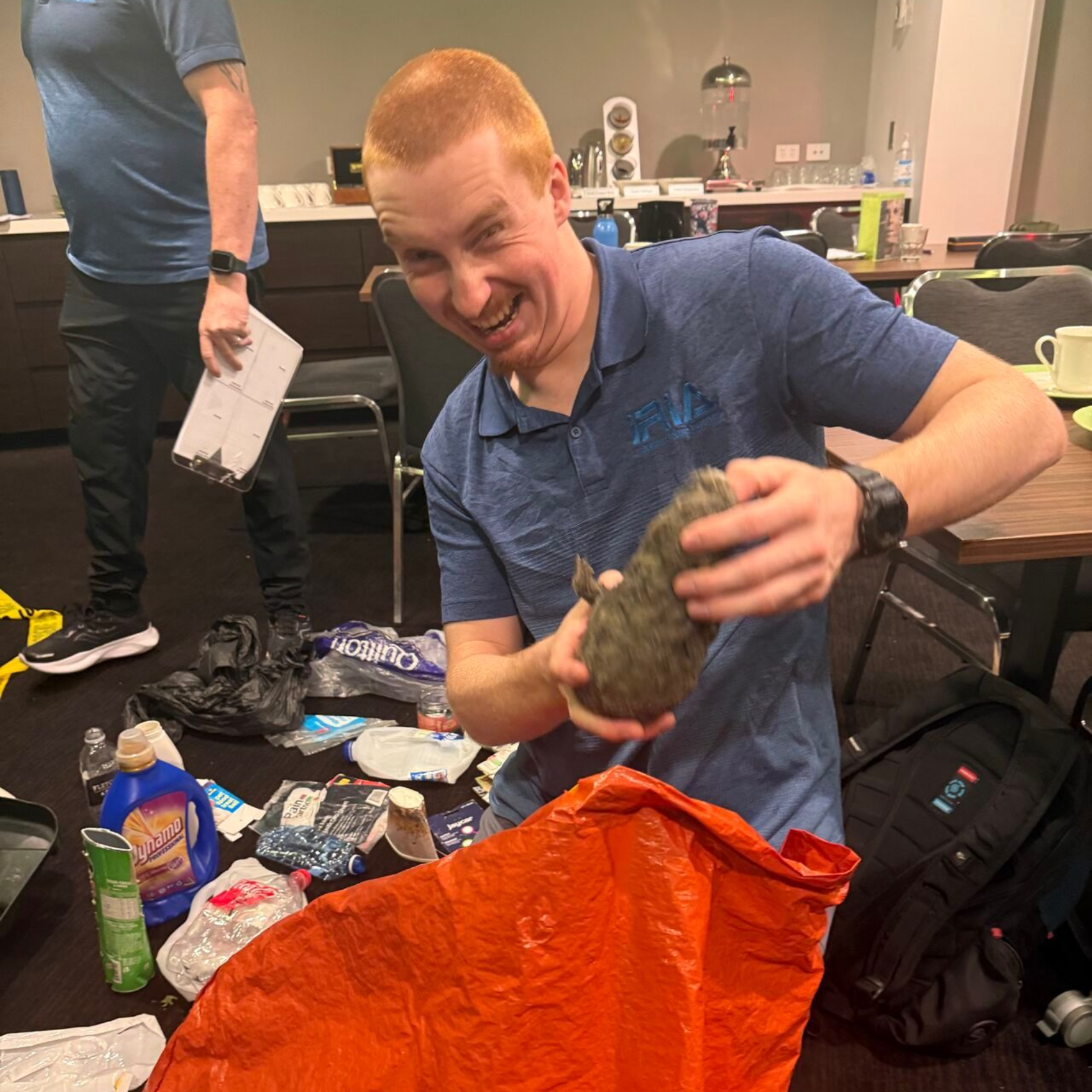 Man with red hair smiles, holding a rock, surrounded by trash. Indoors.