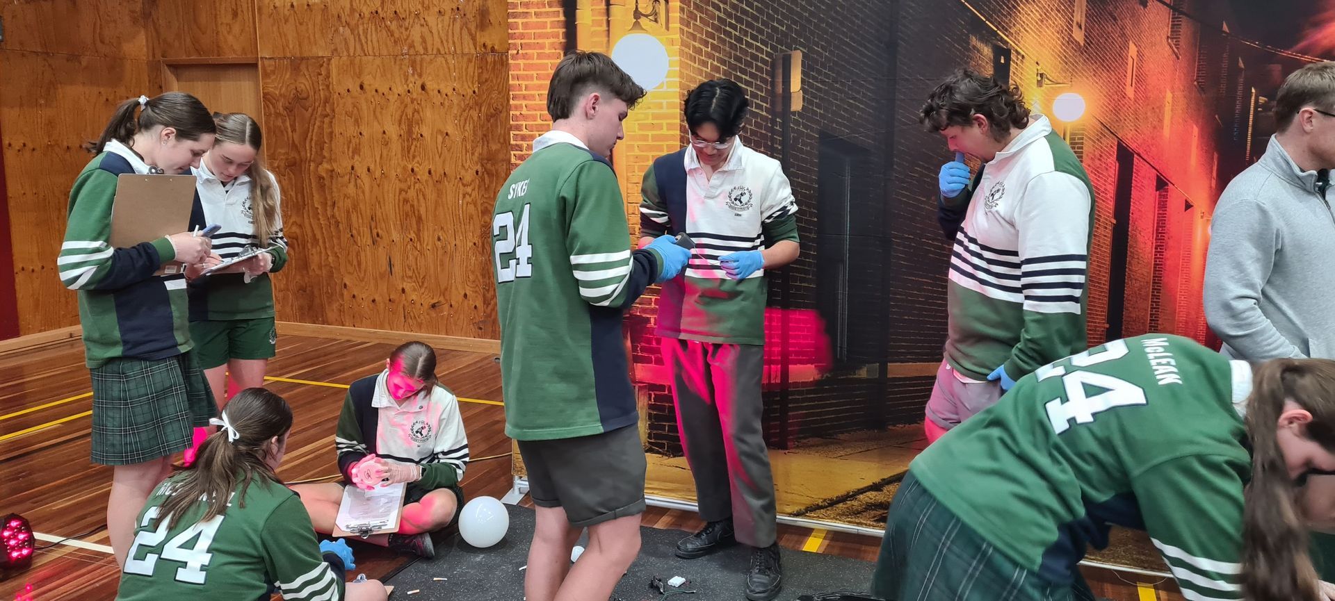 Students in green jerseys work together, some writing, some looking at objects, in a wood-paneled room.