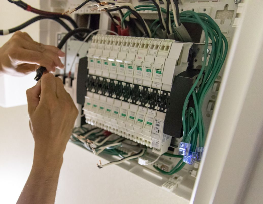 Hands working on an open electrical panel with wires and circuit breakers.