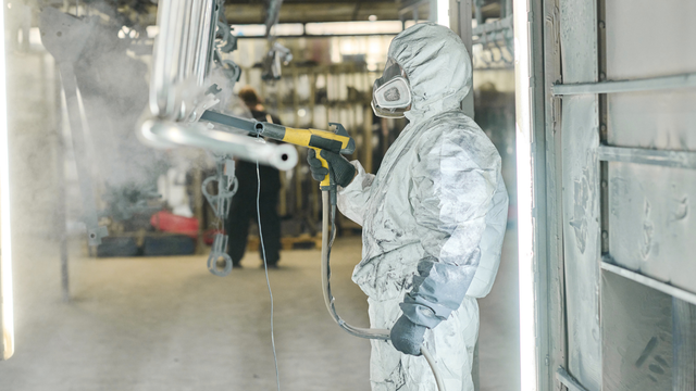 A man in a protective suit is painting a metal object in a factory.