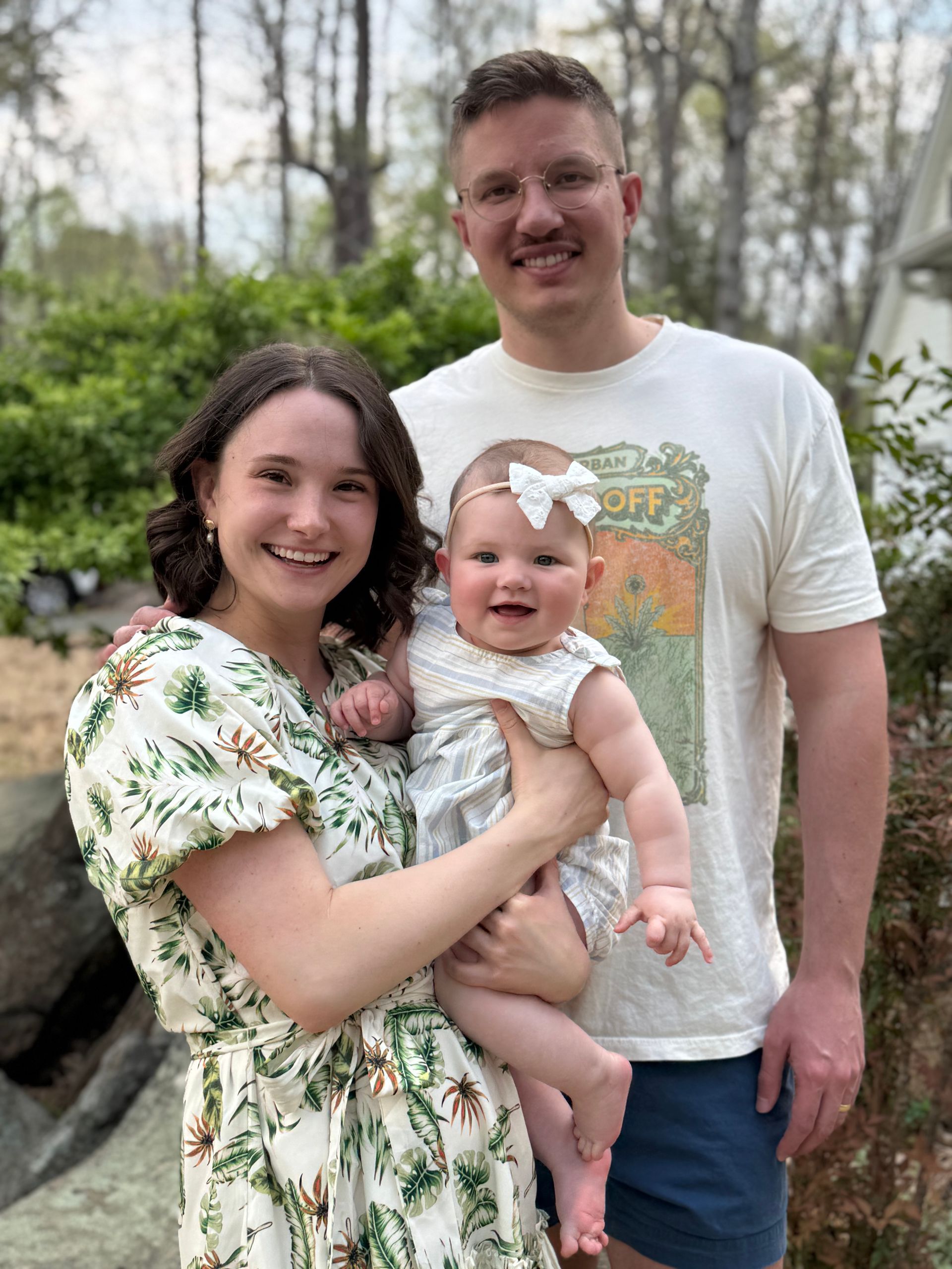 Smiling family of three outdoors, with a woman holding a baby and a man standing behind them.