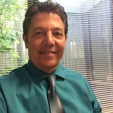 Man in teal shirt and tie, smiling in front of a window with blinds.