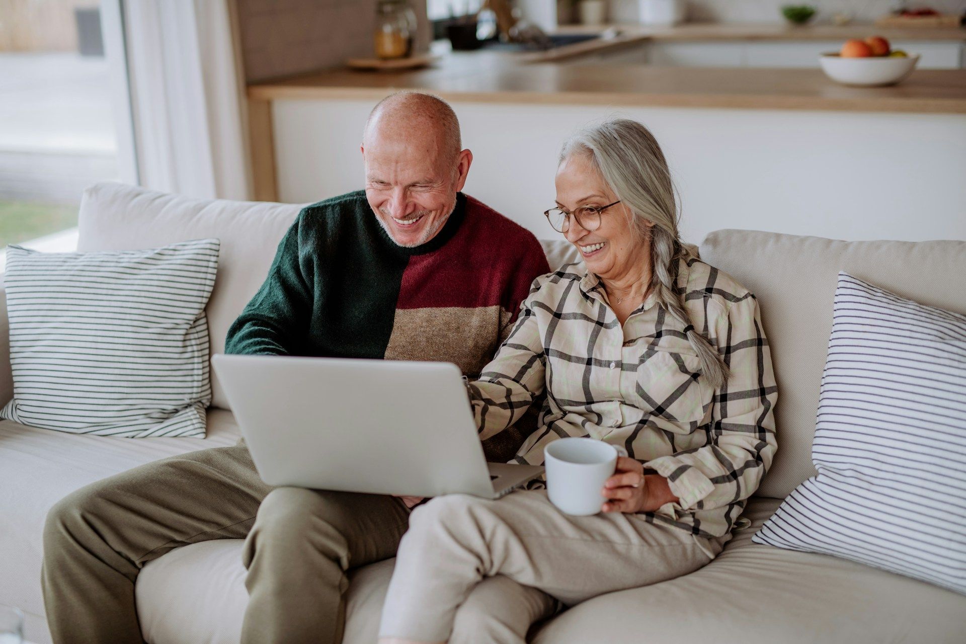 Elderly couple sits on a couch, looking at a laptop and smiling. Indoors, light setting.