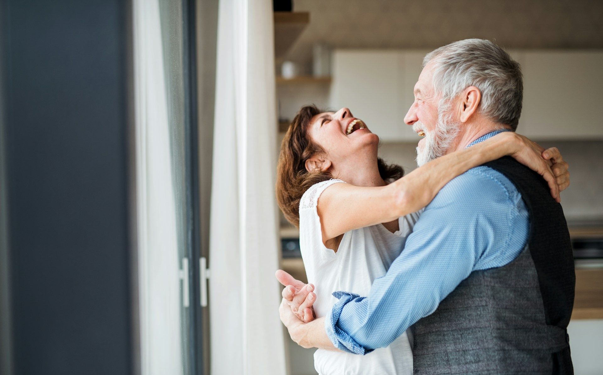 Older couple dancing, laughing, embracing in their home. Man in vest, woman in white shirt.