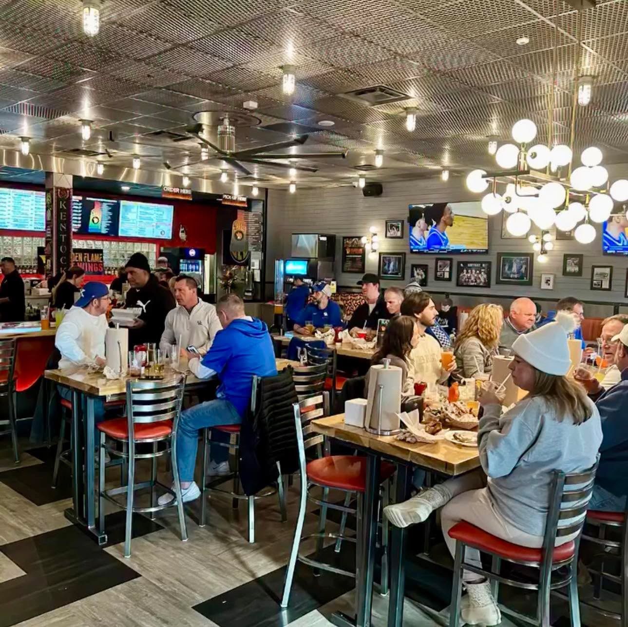 Patrons dine at tables in a lively, well-lit restaurant with a modern metal ceiling and round hanging globe lights.