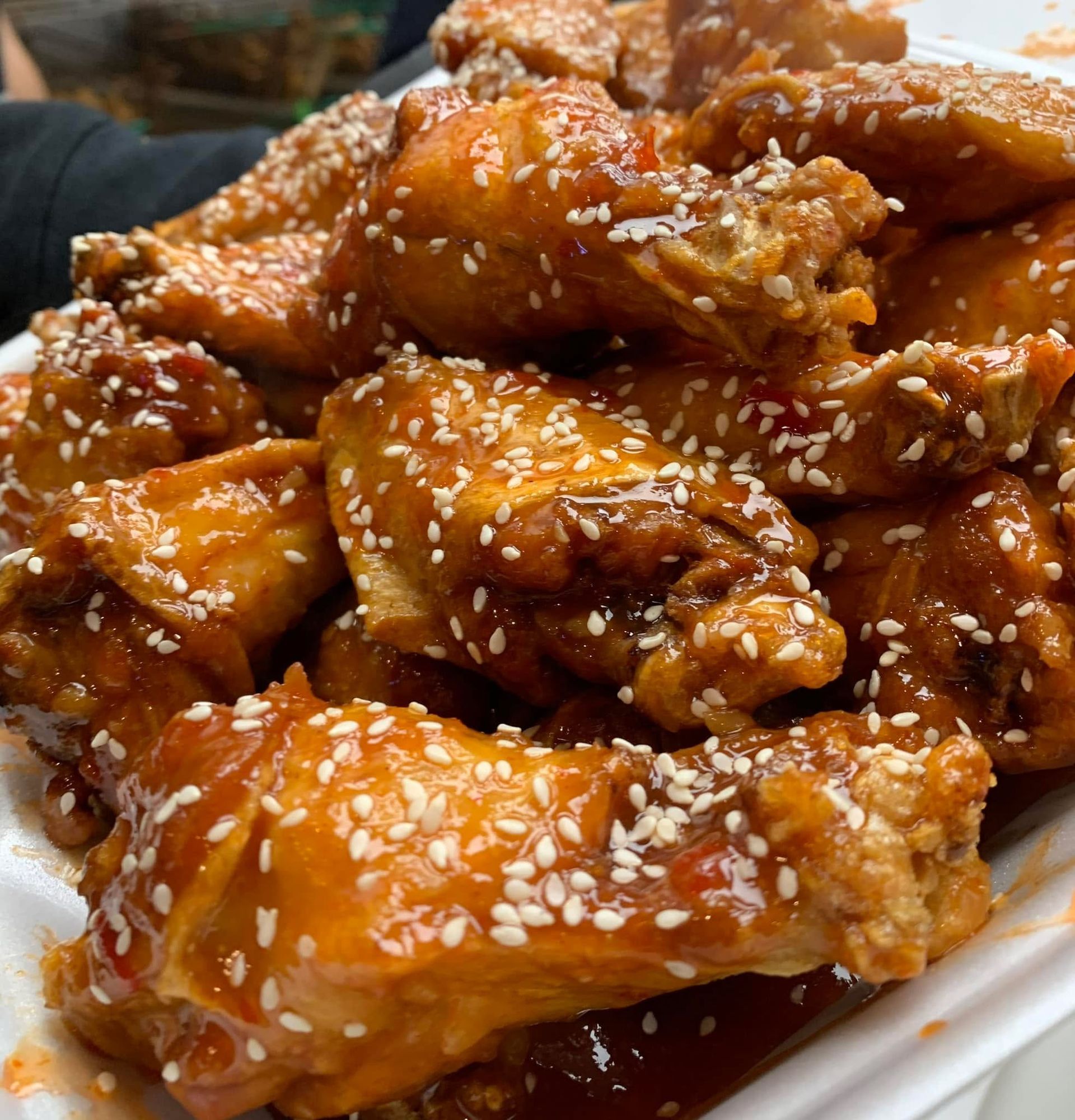 A close-up of several golden-brown chicken wings coated in a shiny, red glaze and sprinkled with white sesame seeds.