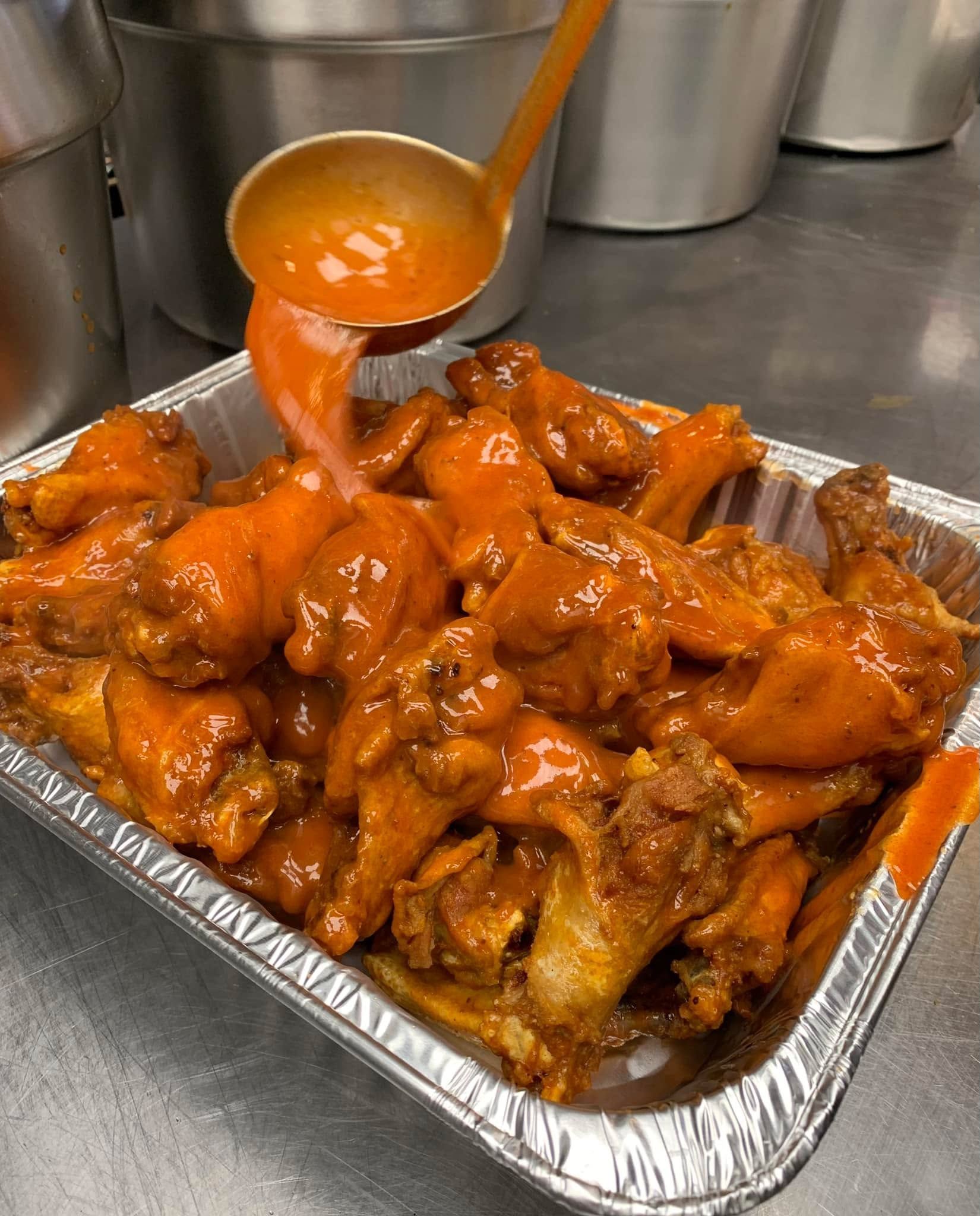A metal ladle pours bright orange buffalo sauce over a tray of fried chicken wings in a commercial kitchen.