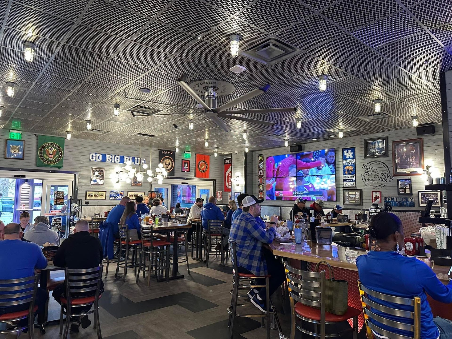 Patrons sit at tables and a bar in a sports bar with a metallic, patterned ceiling, large TV, and blue decor.
