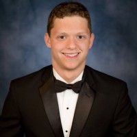 A smiling individual wearing a formal black tuxedo with a bowtie against a mottled blue studio background.